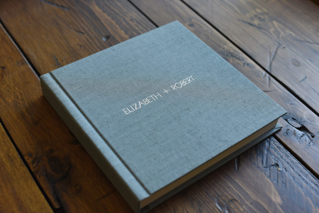 A grey linen wedding album sitting closed on a wooden table, with the names Elizabeth and Robert on the cover.